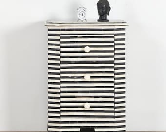 May include: A black and white striped three-drawer nightstand with round white knobs. A small silver alarm clock and a black Buddha head statue sit on top of the nightstand. The nightstand is made of wood.
