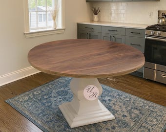 May include: A round wooden dining table with a light brown top and a white pedestal base. The table is set on a blue patterned rug in a kitchen setting. The table top is approximately 48 inches in diameter.