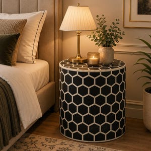 May include: A cylindrical side table featuring a black and white geometric honeycomb pattern. The table surface holds a brass-toned table lamp with a pleated shade, a small glass candle holder with a lit candle, and a potted green plant.