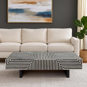 May include: A rectangular coffee table with a black and white geometric pattern. The table has a series of black and white lines arranged in a repeating pattern. The table is supported by black legs. The table is in front of a cream-colored sofa.