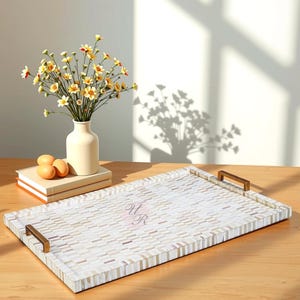 May include: Rectangular serving tray with a mosaic tile design in neutral tones. The tray has brass-colored handles and the initials "UR" in a pink script. A vase of yellow flowers and a stack of books are on the table.