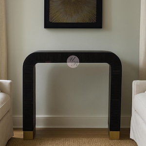 May include: A black console table with a curved design and gold-colored accents on the feet. The table is made of a dark wood with a textured surface. A piece of art hangs above the table.