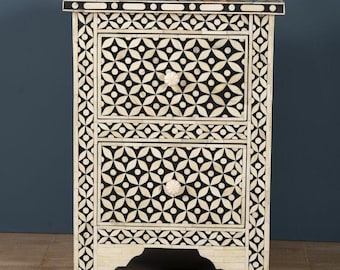 May include: A two-drawer nightstand with a black and white geometric pattern. The nightstand has a decorative border and a small, terracotta-colored bowl on top. The drawers have floral-shaped knobs. The nightstand is made of wood.