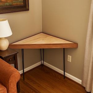 May include: A triangular corner table with a light wood top and a dark wood trim. The table is supported by three black metal hairpin legs. The table is approximately 30 inches wide at its longest point.