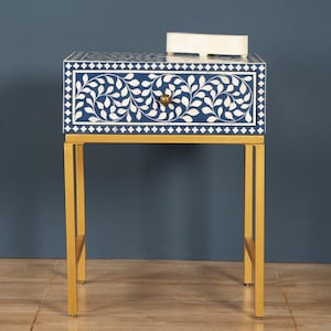 May include: A navy blue nightstand with a white floral inlay design. The drawer has a gold-colored knob. The table is supported by gold-colored metal legs. A small, rectangular, off-white object rests on the top surface.