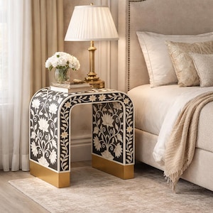 May include: A black and white floral patterned side table with gold accents. The table has a curved design and is topped with a lamp, flowers, and books. The table is next to a bed with a beige headboard.