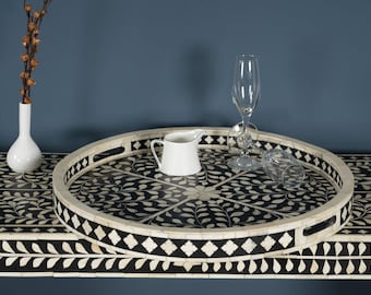 May include: A decorative serving tray with a black and white floral design, a white creamer, and two champagne flutes. A white vase with dried stems is on the left. The tray is on a black and white table.