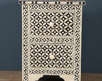 May include: A two-drawer nightstand with a black and white geometric pattern. The nightstand has a cream-colored base and is topped with a small, brown, terracotta bowl. The drawers have white, flower-shaped knobs.