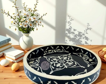 May include: A round decorative tray with a black interior and white floral design. The tray has a navy blue border with white accents. A white vase with white flowers sits to the left of the tray.