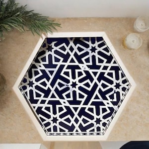 May include: A hexagonal decorative tray with a white geometric pattern over a dark blue background. The tray is set on a beige surface, with a sprig of greenery and candles in the background.