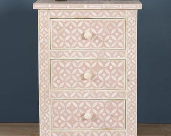 May include: A pink and white patterned nightstand with three drawers. The nightstand has a floral design and white knobs. A small Statue of Liberty and a white alarm clock sit on top of the nightstand.