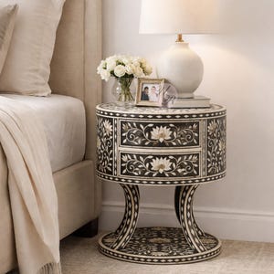 May include: A black and white round nightstand with two drawers, featuring floral inlay designs. A white lamp, a vase of white flowers, and a framed photo sit on top. The nightstand has a curved base and is next to a bed.