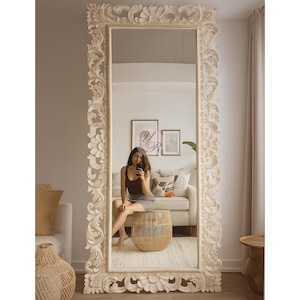 May include: A large, ornate, full-length mirror with a cream-colored, carved wooden frame. The mirror reflects a room with a sofa, artwork, and a person sitting on a wooden stool. The frame features floral and scrollwork details.