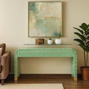 May include: A green and white patterned console table with a glass top, gold-colored feet, and a matching framed painting above. Two small white pots with plants and a decorative box sit on the glass shelf. A potted plant is on the right.