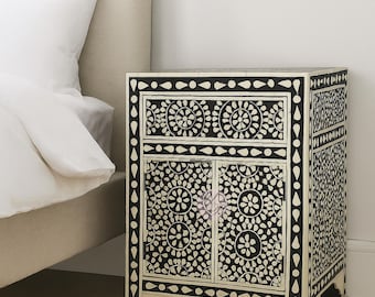 May include: Black and white floral patterned nightstand with two doors and a drawer. The nightstand has a cream-colored base and is placed next to a bed with white bedding. The nightstand is approximately 24 inches tall.