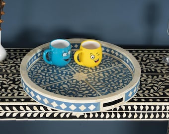 May include: A round decorative tray with a blue and white floral design, holding two ceramic mugs. One mug is blue with a smiling face, and the other is yellow with a winking face. The tray has a light blue border.