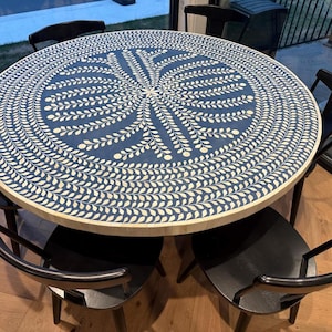 May include: A round dining table with a blue surface featuring an intricate white leaf pattern radiating from the center. The table edge is finished with a light-colored material, and it is surrounded by several black chairs with curved backs.