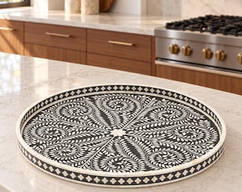 May include: A round tray featuring a black surface with an intricate white floral and leaf pattern radiating from the center. The tray has a raised edge decorated with a repeating white diamond pattern against a black background, creating a contrast.