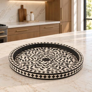 May include: A round tray featuring a black and white floral pattern on the interior base and a repeating cross-shaped motif along the outer rim. The tray has a low, raised edge and a flat bottom, displaying a high-contrast design throughout.