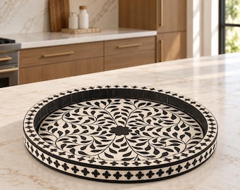 May include: A round tray featuring a black and white floral pattern on the interior base and a repeating cross-shaped motif along the outer rim. The tray has a low, raised edge and a flat bottom, displaying a high-contrast design throughout.
