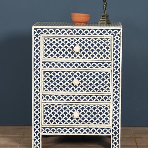 May include: A three-drawer nightstand with a navy blue and white scallop pattern. The nightstand has white knobs and is topped with a small terracotta bowl and a Statue of Liberty figurine. The nightstand is approximately 30 inches tall.