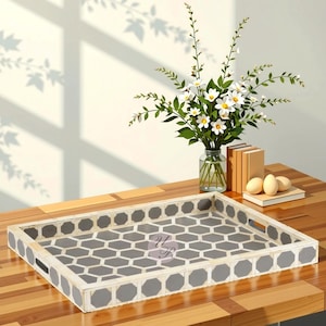 May include: A rectangular decorative tray with a gray and white geometric pattern. The tray has a white border with gray octagonal accents. A vase of white flowers and books are on a wooden table.