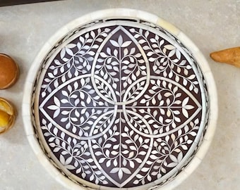 May include: Round decorative tray with a white rim and a dark brown center. The center features a white floral leaf pattern. The tray is likely made of wood and is suitable for serving or display.