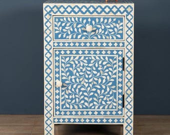 May include: A blue and white floral patterned nightstand with a drawer and a cabinet door. The nightstand has a white border with a geometric design. A small, brown bowl sits on top. The nightstand is approximately 24 inches tall.