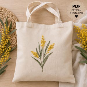 May include: A cream-coloured canvas tote bag featuring an embroidered design of yellow mimosa flowers with green leaves. The bag has two top handles and a circular badge in the top right corner with the text PDF PATTERN DOWNLOAD.