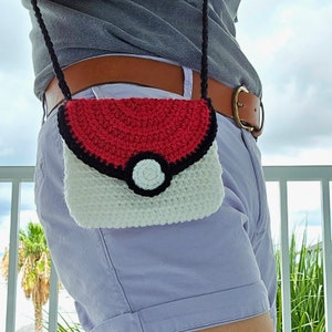 May include: A crochet crossbody bag in a red, black, and white color scheme. The bag features a circular design with a black border and a white button detail. The strap is black.