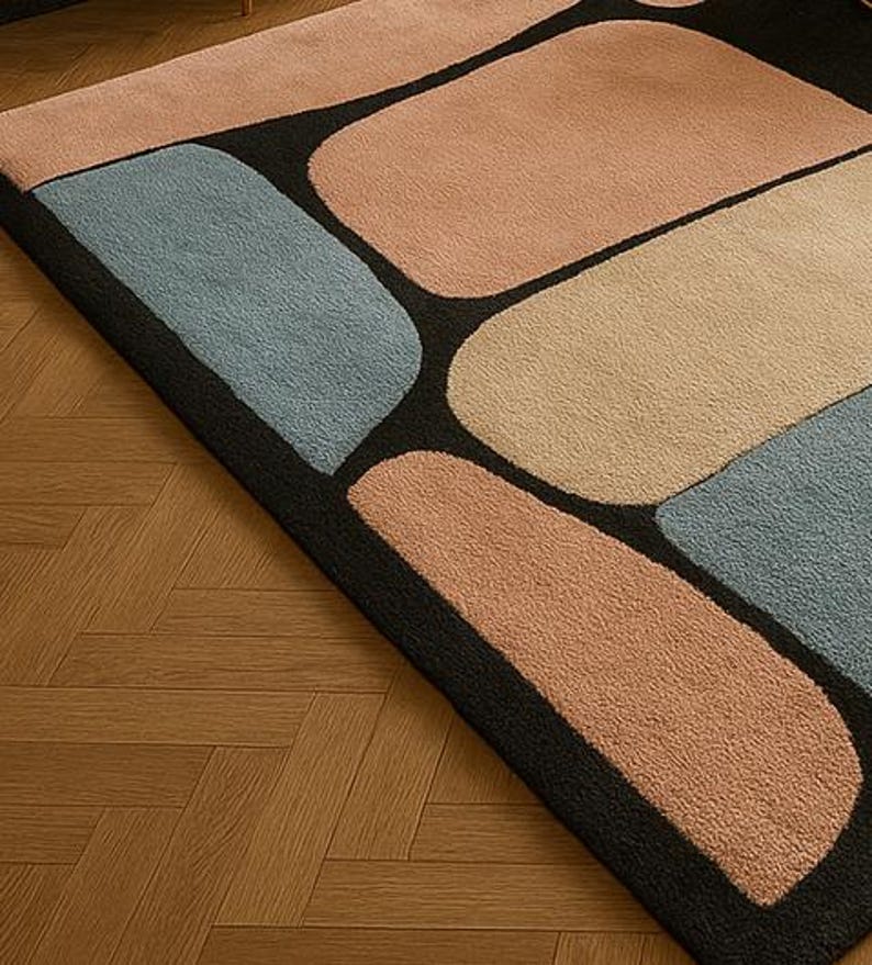 May include: A modern area rug with a geometric design. The rug features large, rounded rectangular shapes in shades of peach, beige, and blue, set against a black background. The rug is placed on a wooden floor.