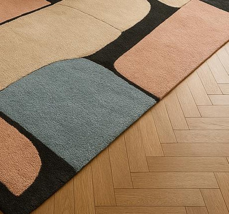 May include: A modern area rug with a geometric design. The rug features large, irregular shapes in shades of beige, peach, and blue, separated by black lines. The rug is placed on a wooden floor with a herringbone pattern.