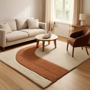 May include: A cream-colored area rug with a large, curved, rust-colored design. The rug is in a living room setting with a beige sofa, a round wooden coffee table, and a rust-colored armchair. The room has hardwood floors and a window with sheer curtains.