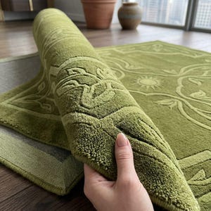 May include: A sage green area rug featuring a raised, embossed floral and sunburst pattern. The rug is partially rolled, revealing a thick, tufted texture and a finished border. The design consists of symmetrical botanical motifs and a central sun.