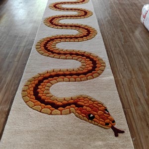 May include: A long, rectangular runner rug featuring a cream-coloured background with a large, winding snake design. The snake is rendered in shades of orange, gold, and dark brown, with a textured, tufted appearance and a forked tongue at the head.