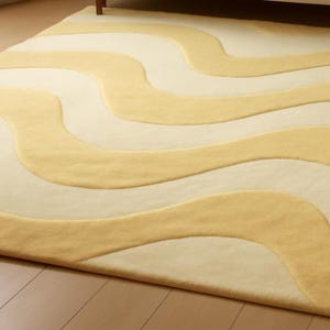 A hand-tufted wool rug on a wooden floor featuring an abstract wavy pattern in alternating bands of butter yellow and cream.
