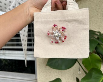 Könnte beinhalten: Eine kleine, beige Canvas-Tragetasche mit einer aufgenähten Blumenverzierung aus Perlen. Die Blume hat rote und klare Perlen um eine zentrale Perle. Die Tasche hat eine rechteckige Form mit einem Griff.