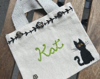 Könnte beinhalten: Kleine, cremefarbene Canvas-Tragetasche mit kurzem Henkel. Die Tasche zeigt den Namen "Kat" in Grün gestickt, eine schwarze Katzenillustration und dekorative schwarze Nähte mit floralen Akzenten und dekorativen Knöpfen.