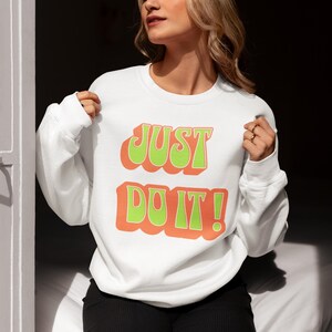 Puede incluir: Sudadera blanca con el texto "JUST DO IT!" en letras verdes y naranjas.