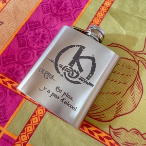 May include: A silver flask with a black inscription that reads "Ouais... En plus, y a pas d'alcool." The flask has a circular design with a stylized symbol.
