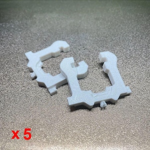 Op de afbeelding: Twee witte, plastic, sleutelvormige clips worden getoond op een getextureerd oppervlak. Het nummer "x 5" is in het rood in de linkerbenedenhoek gedrukt. De clips hebben een uniek ontwerp met een gebogen bovenkant en een gekerfde basis.