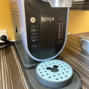 Op de afbeelding: Een zwarte en zilveren Ninja-blender met knoppen voor verschillende drankopties. Een lichtblauwe, ronde onderzetter met een Mickey Mouse-ontwerp staat ervoor. De tekst "Grille uniquement" is zichtbaar.