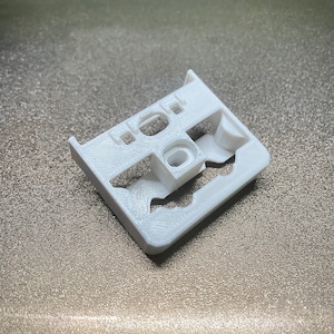 Peut inclure: Pièce en plastique blanc imprimée en 3D de forme rectangulaire avec plusieurs découpes. La pièce a une surface texturée.