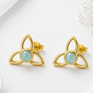 May include: A pair of gold-toned stud earrings featuring a triquetra-inspired openwork design. Each earring centres a smooth, round, pale mint-green cabochon stone set within a polished gold frame that forms three symmetrical, petal-like loops.
