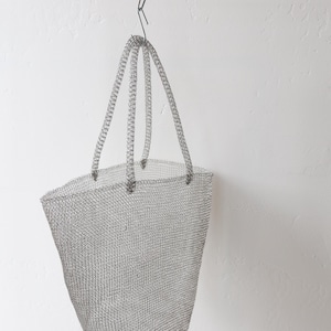 May include: A silver-colored metal mesh tote bag with three handles is suspended from a black metal bar. The bag has a rectangular shape with a wide opening and a textured surface. The background is a plain white wall.