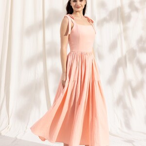 May include: A woman wearing a peach-colored tiered maxi dress with thin straps that tie at the shoulders. The dress has a square neckline and a flowy skirt. She is wearing beige heeled sandals.