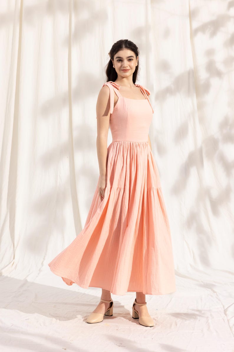 May include: A woman wearing a peach-colored tiered maxi dress with thin straps that tie at the shoulders. The dress has a square neckline and a flowy skirt. She is wearing beige heeled sandals.