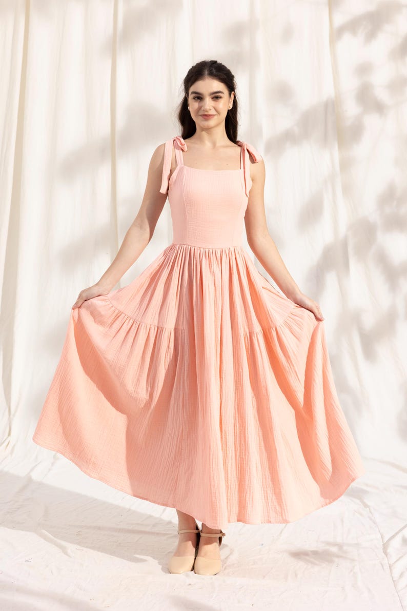May include: A peach-colored tiered maxi dress with thin straps that tie at the shoulders. The dress has a square neckline and a flowy skirt.