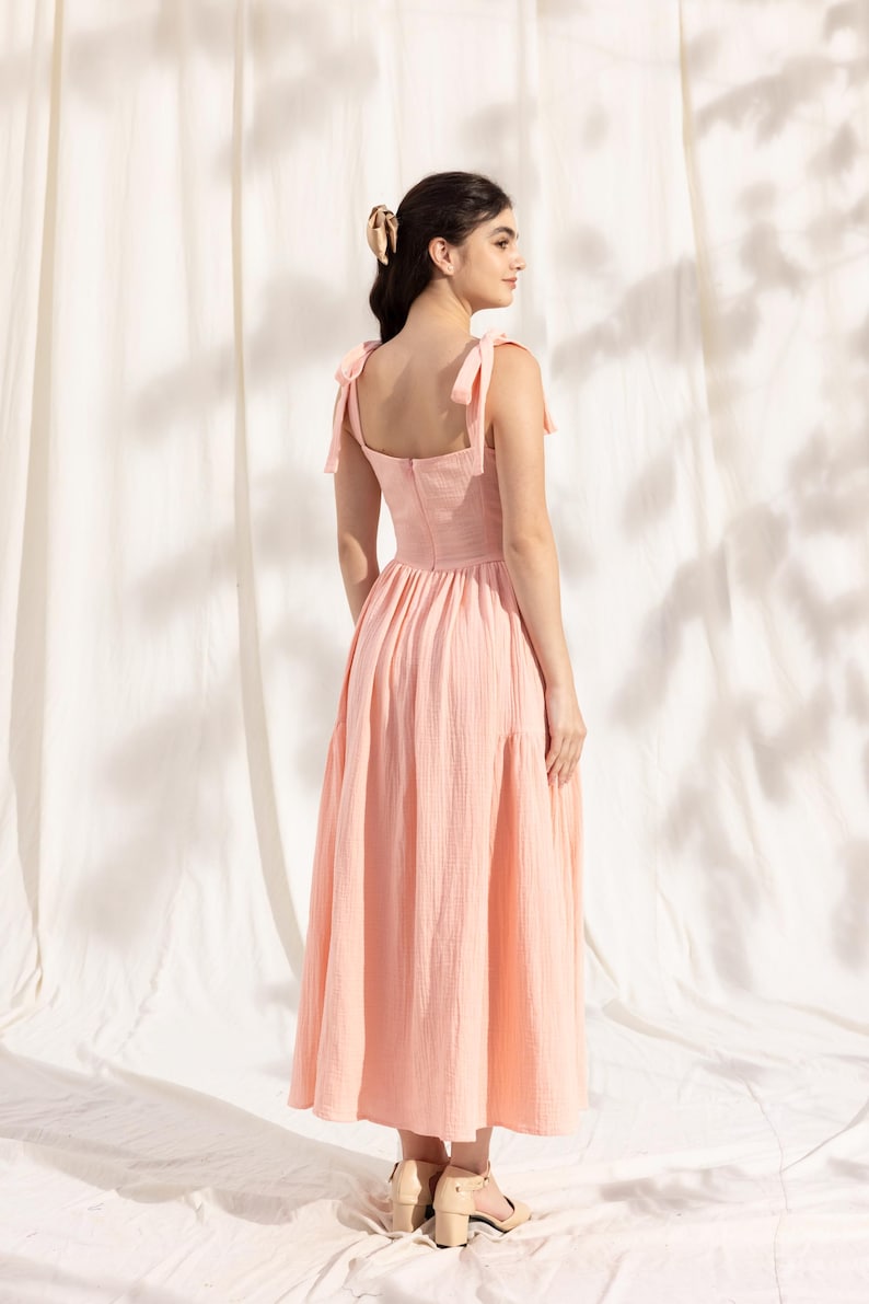 May include: A light pink midi dress with a square neckline and thin straps that tie at the shoulders. The dress has a gathered skirt and a back zipper closure.
