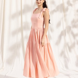May include: A peach-colored tiered maxi dress with thin straps that tie at the shoulders. The dress is made of a lightweight fabric and has a flowy silhouette.